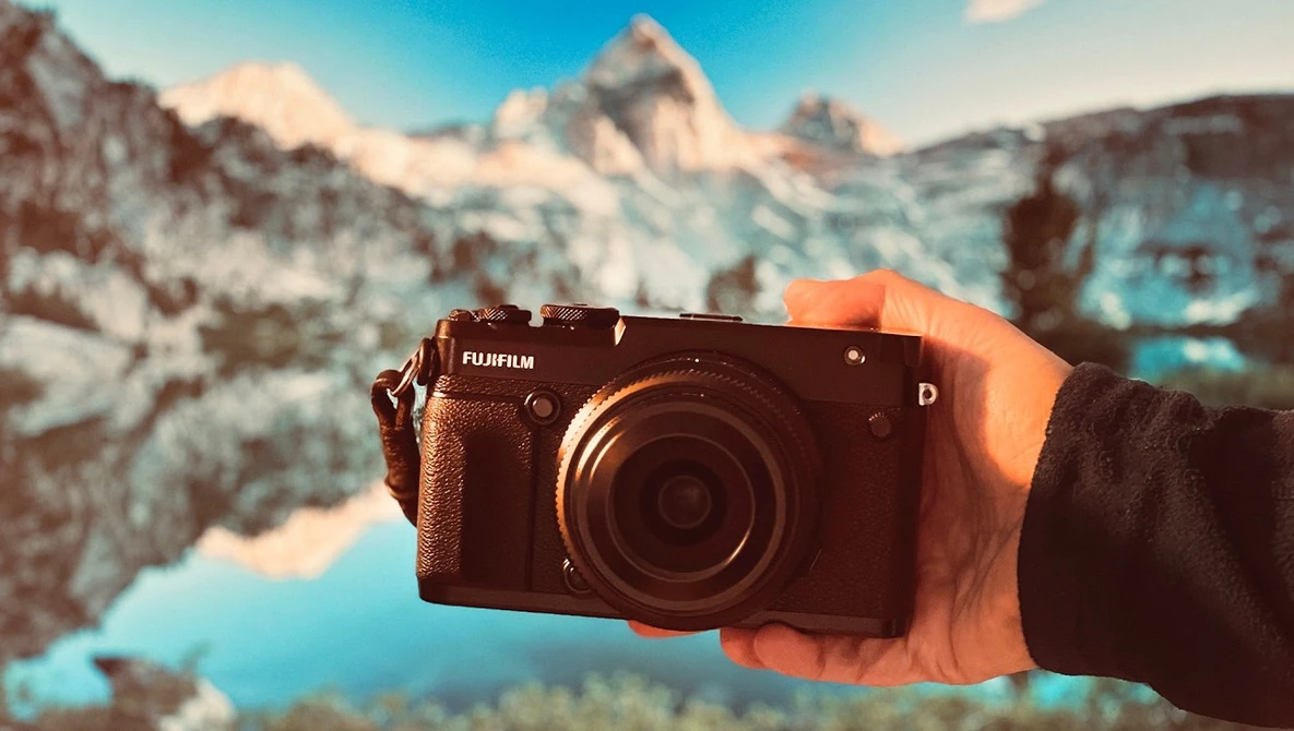 Hand holding a Fujifilm digital camera with snow-capped mountains and a turquoise lake in the background.