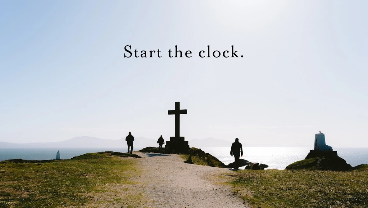 Silhouetted figures stand on a hillside near a large cross overlooking a coastal landscape at dusk.