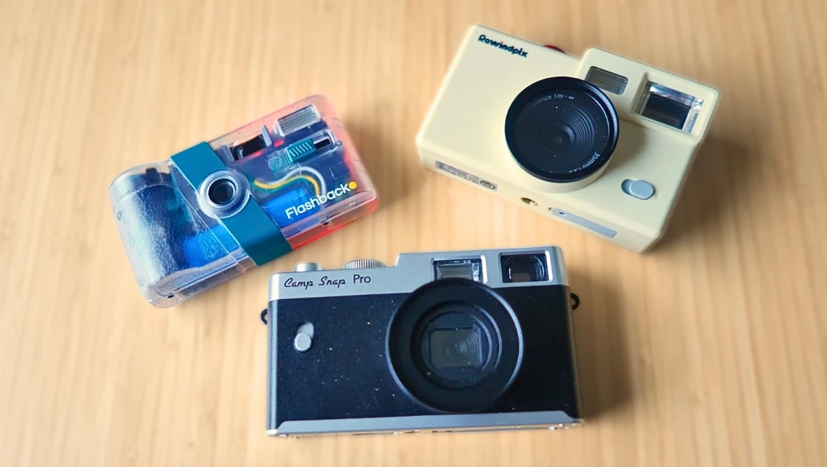 Three instant and film cameras arranged on a wooden surface: a blue disposable camera, a cream-colored instant camera, and a black rangefinder camera.
