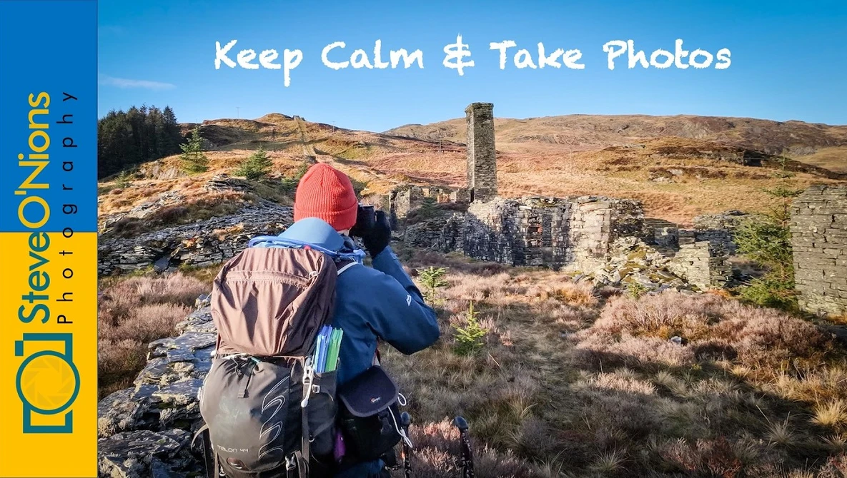 Hiker with red beanie and backpack photographing ruined stone structures in moorland landscape.