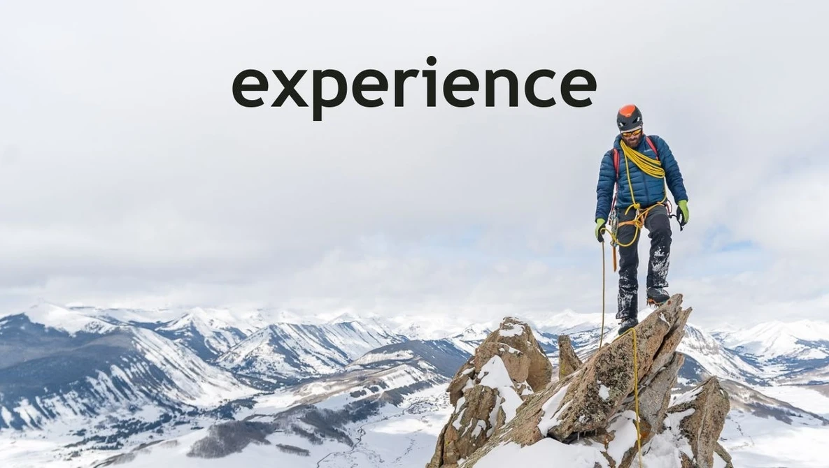 Mountain climber standing on rocky summit overlooking snow-covered alpine landscape.