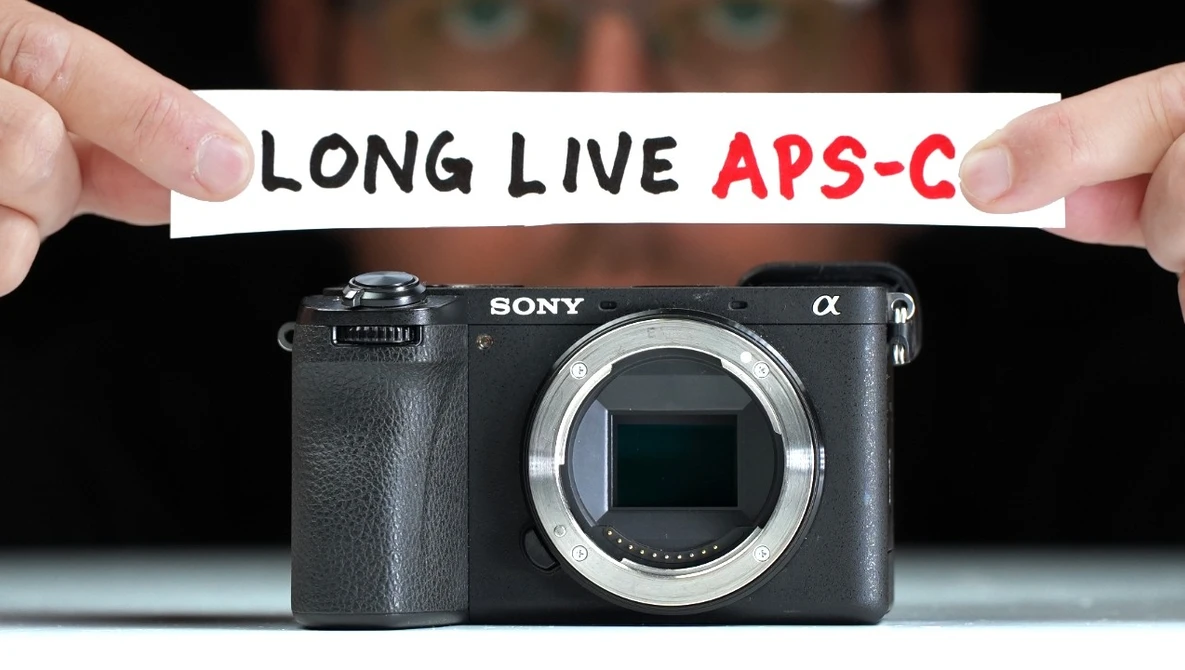 Sony Alpha mirrorless camera body with text banner reading "LONG LIVE APS-C" held above it.