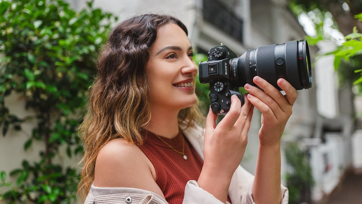 Woman holding a mirrorless camera with telephoto lens, smiling while composing a shot outdoors.