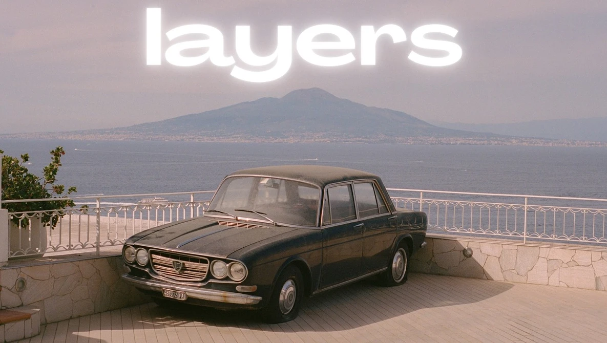 Classic sedan parked on a coastal overlook with a volcanic mountain visible across the water in soft, hazy light.