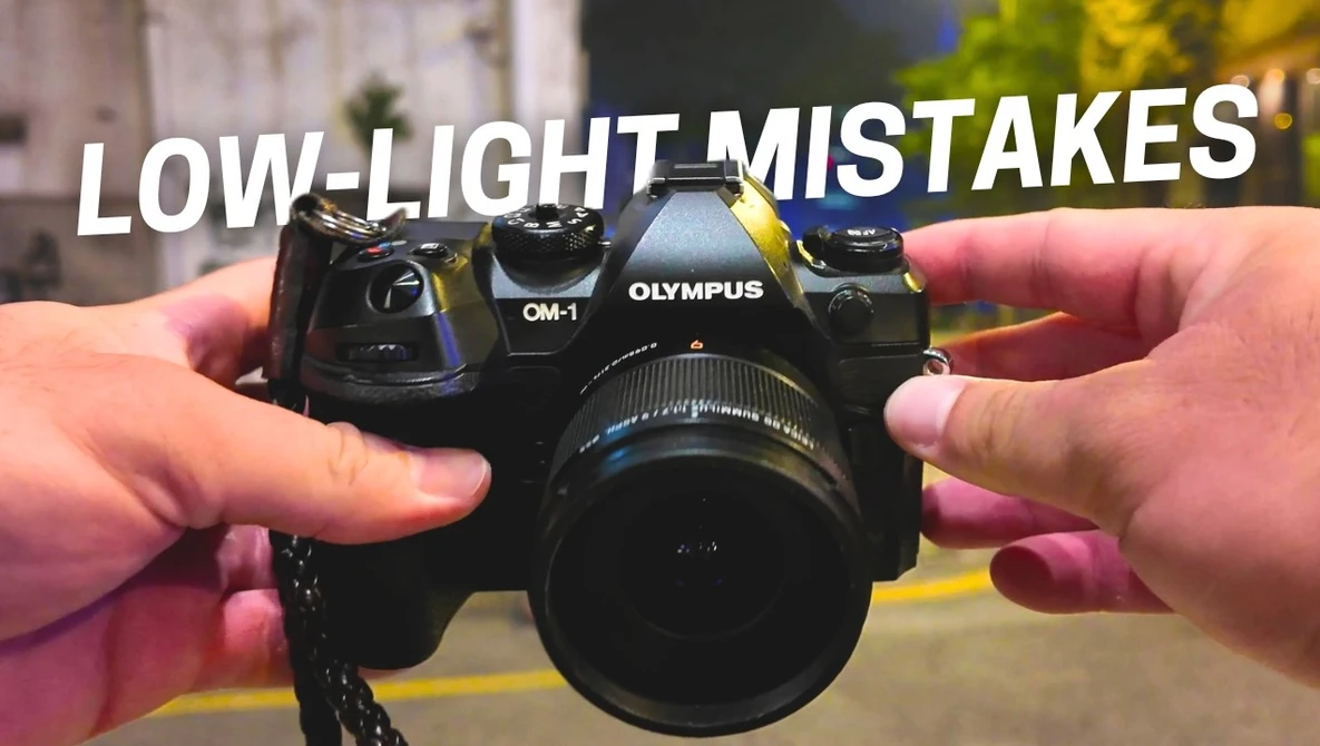 Hands holding an Olympus OM-1 camera with lens against blurred background.