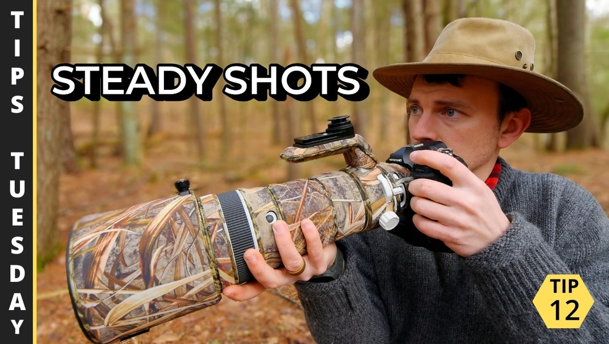 Photographer holding a telephoto lens with camouflage covering while aiming a camera with mounted scope in a forest setting.
