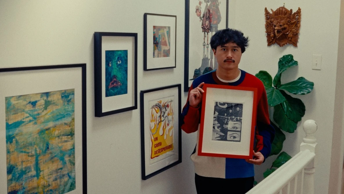 Artist holding a framed black and white print in a gallery space surrounded by various artworks.