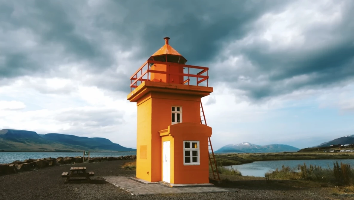 Bright orange and yellow lighthouse structure standing alone on dark sand with mountains and water visible in the background under stormy skies.