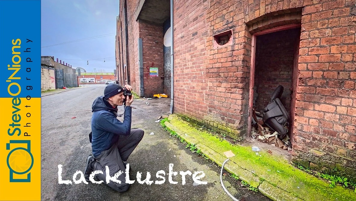 Photographer crouching to compose a shot of a moss-covered ground strip against weathered brick industrial building.
