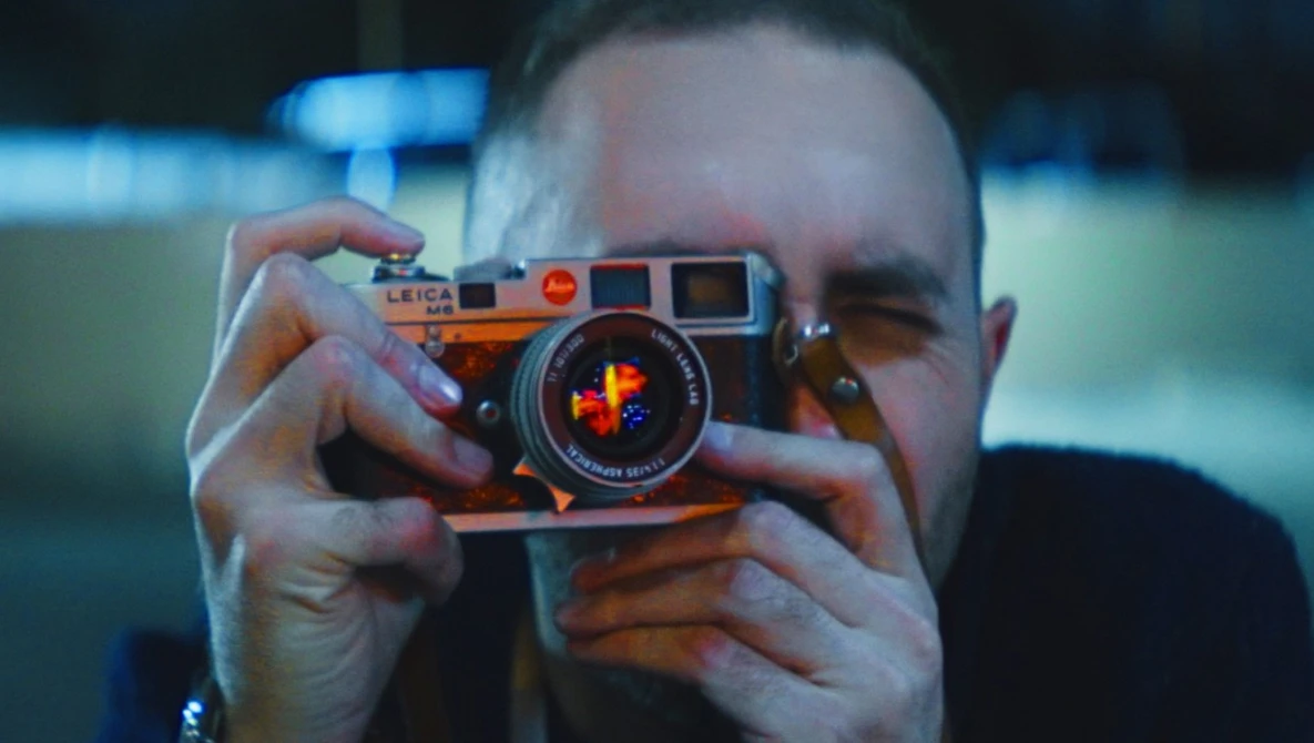 Photographer holding an orange rangefinder camera up to their face, obscuring their head while composing a shot.