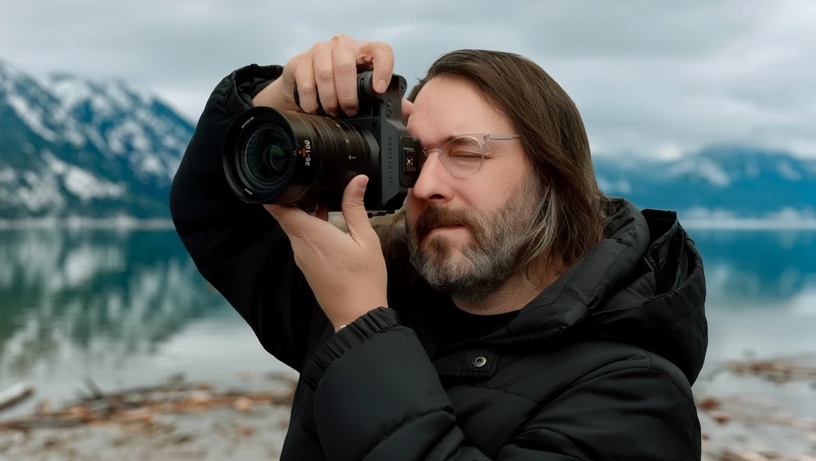 Photographer holding a DSLR camera to his eye while composing a shot of a mountain lake landscape.