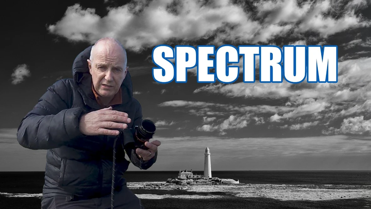 Man in dark jacket holding camera at coastal lighthouse location with dramatic sky.