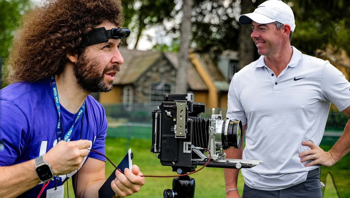 Filmmaker with camera on tripod discussing with golfer on outdoor course.