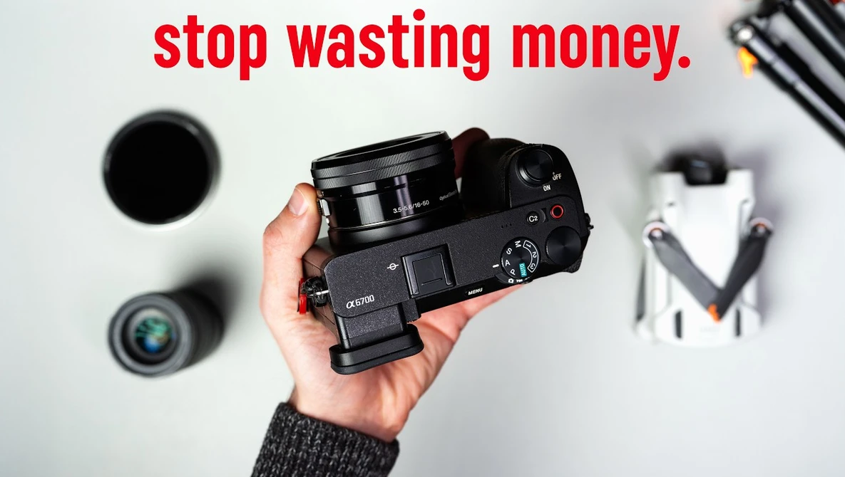 Hand holding a compact mirrorless camera with various photography equipment displayed on a white surface.