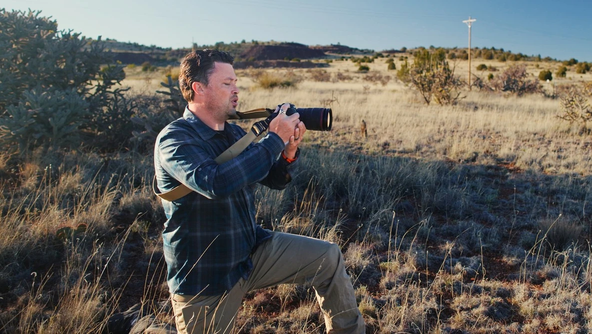 Man holding binoculars to his eyes while crouching in dry grassland.