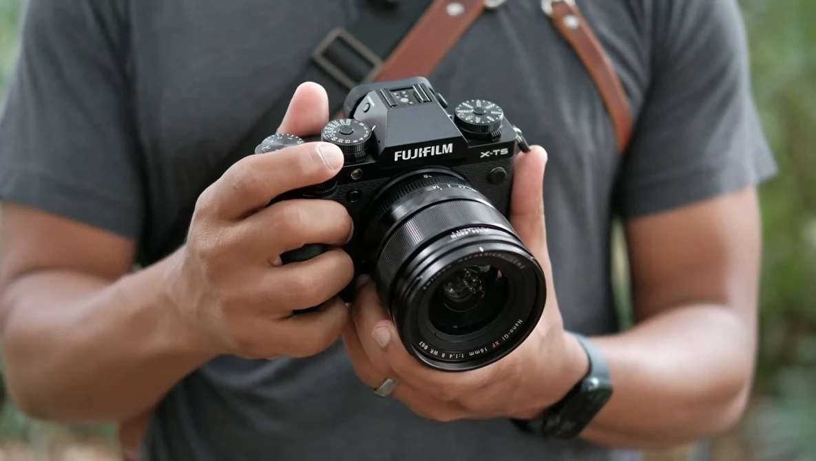 Photographer holding a Fujifilm X-T5 camera with attached lens, wearing a gray shirt and brown camera strap.