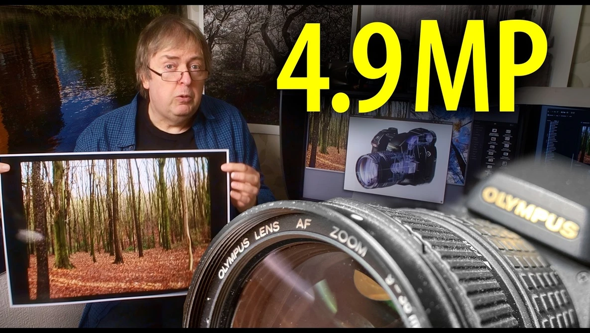Photographer holding printed forest landscape photo next to Olympus lens and camera equipment on desk.