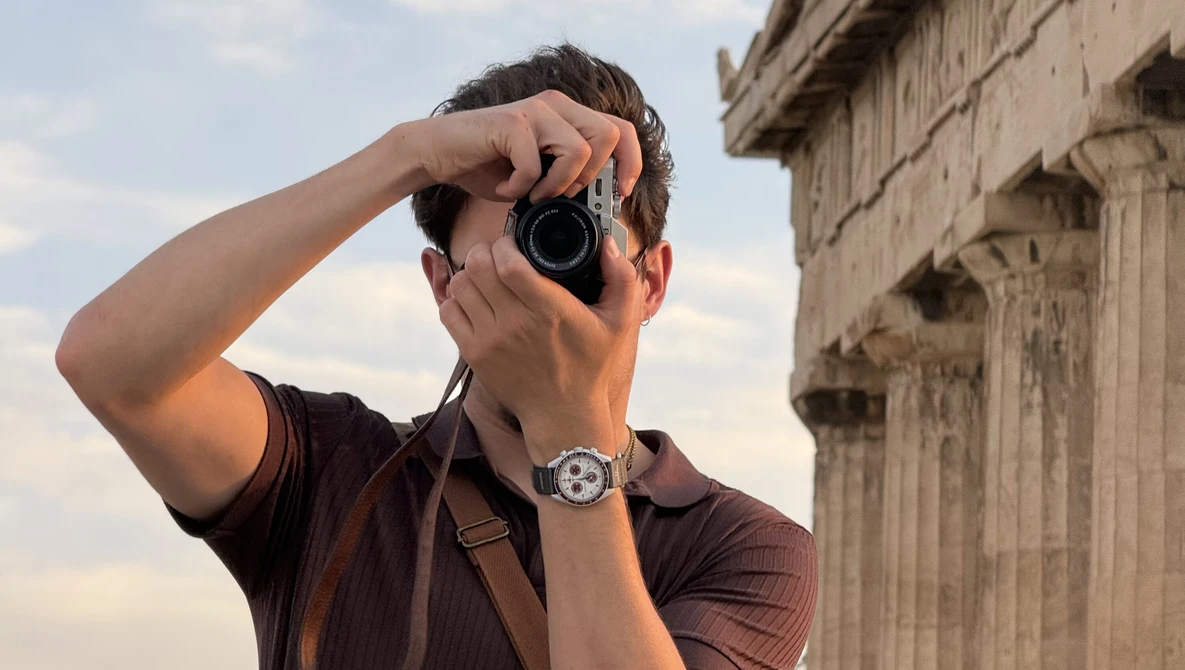 Photographer holding a rangefinder camera to his eye while standing in front of classical stone columns.