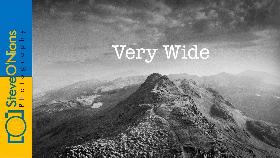 Black and white mountain landscape with a prominent peaked ridge and distant peaks under dramatic cloudy sky.
