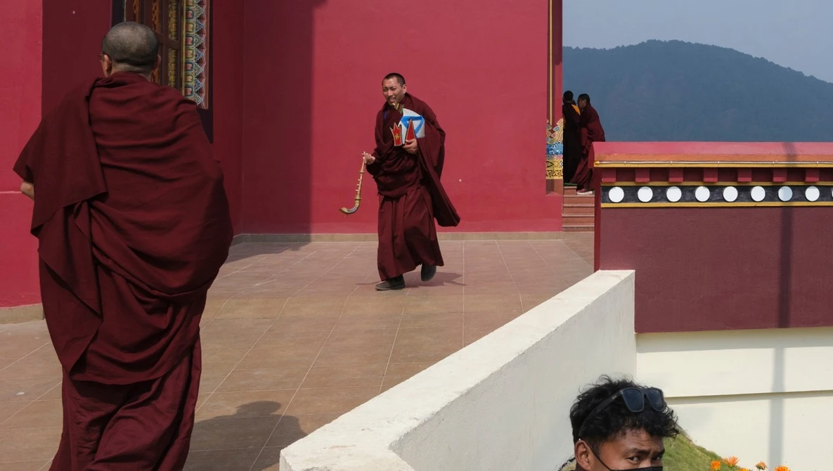 Buddhist monks in maroon robes walking across a courtyard with deep red walls and mountain backdrop
