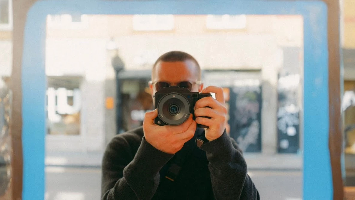 Photographer holding a mirrorless camera up to their face, viewed through a blue-framed mirror or reflective surface.