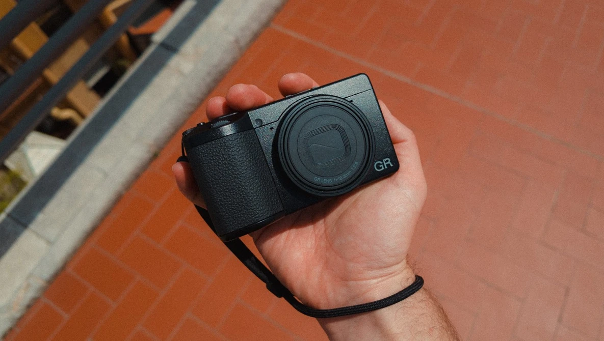 Ricoh GR compact camera held in hand against brick background