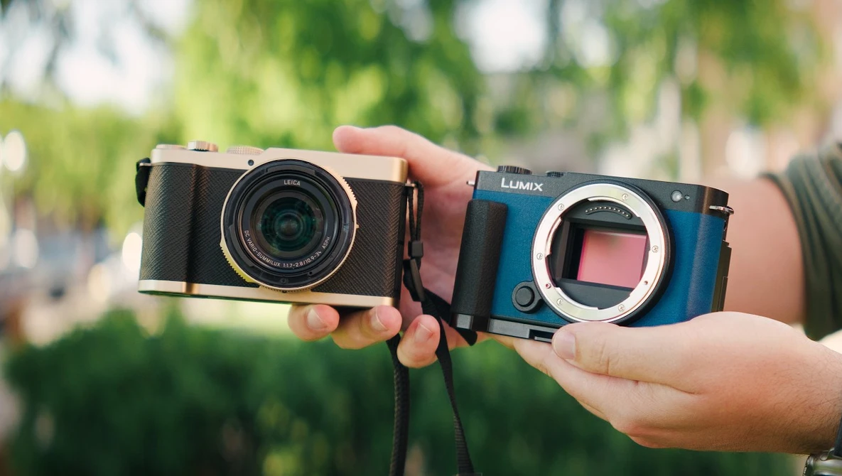 Hands holding a black rangefinder camera and blue Lumix mirrorless camera body side by side outdoors