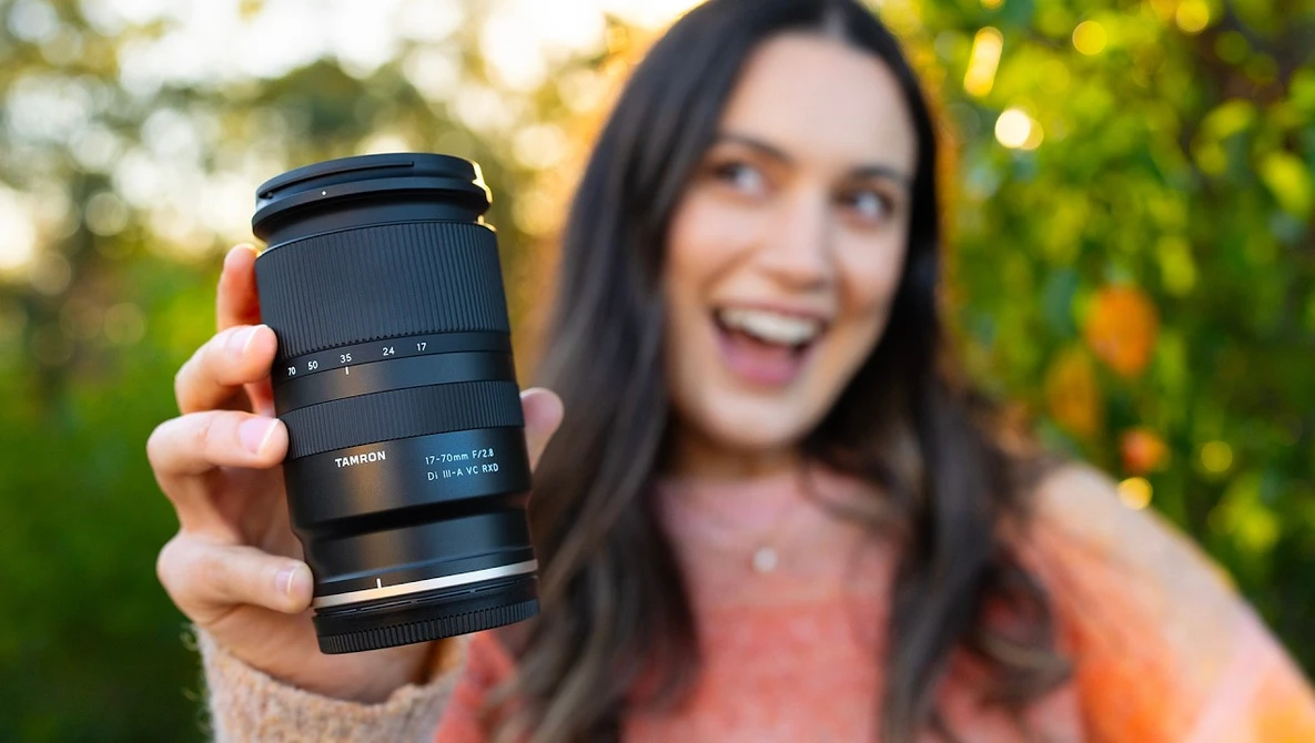 Woman holding a Tamron telephoto lens toward camera in outdoor setting