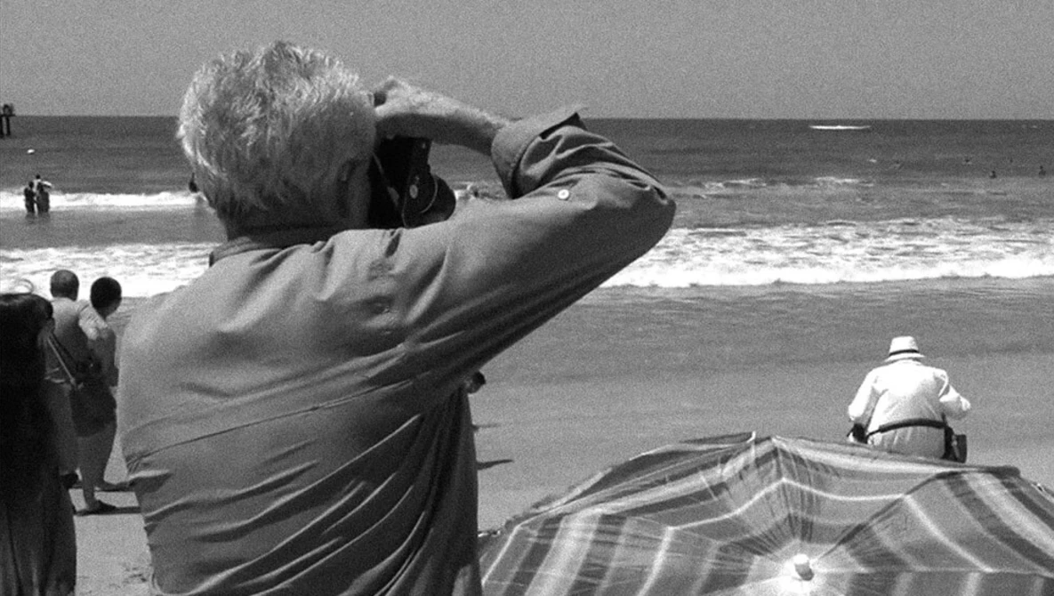 Older man with white hair photographing the ocean from a beach, holding camera to his face