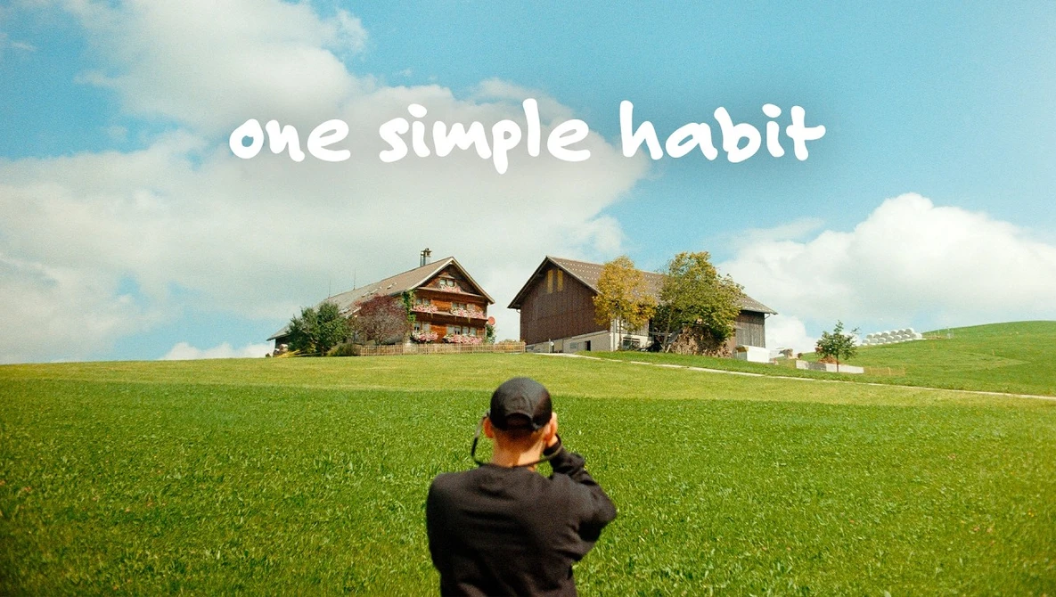 Person photographing rural farmhouses across an open green field under blue sky with text overlay