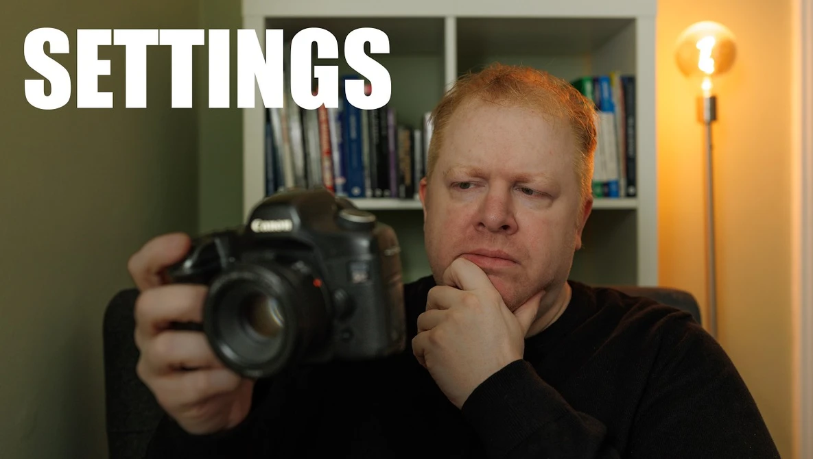 Photographer in black shirt holding Canon DSLR camera with thoughtful expression, hand on chin