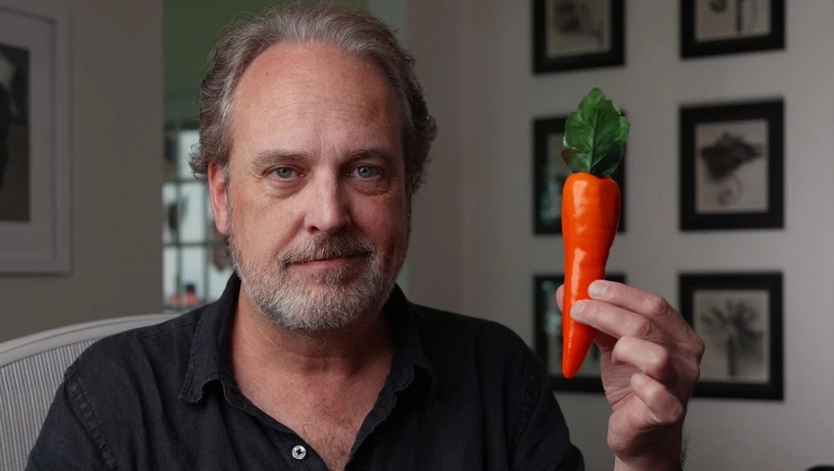 Man holding a fresh carrot with green leafy top in an indoor studio setting