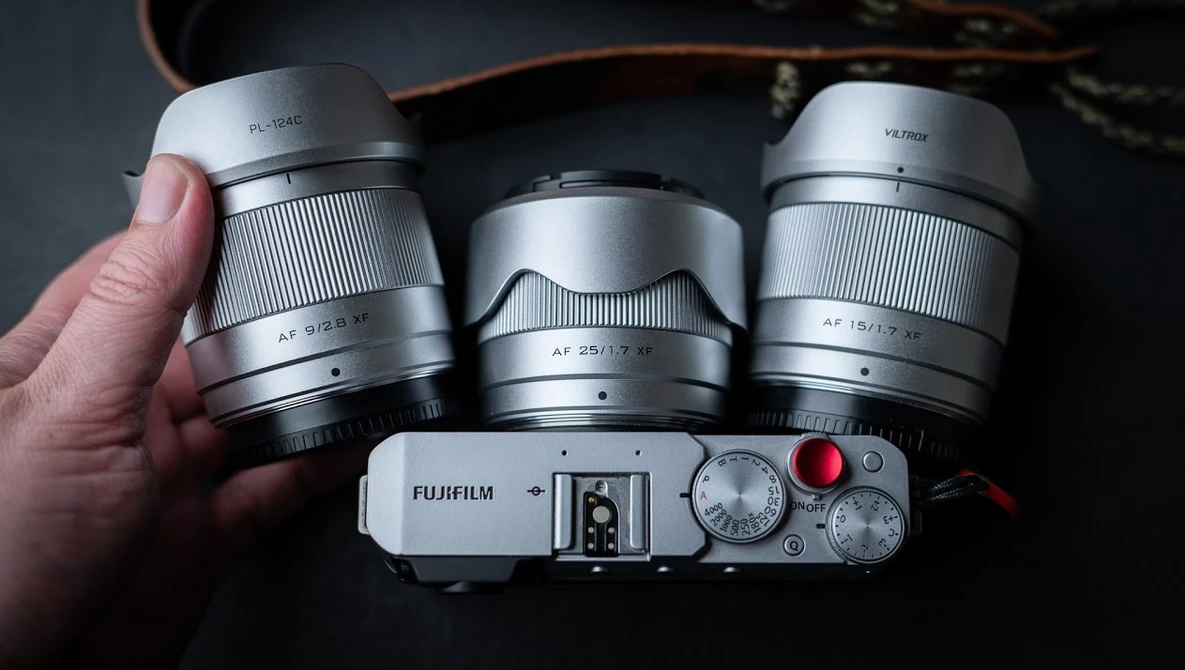 Hand holding silver Fujifilm mirrorless camera with three interchangeable lenses displayed