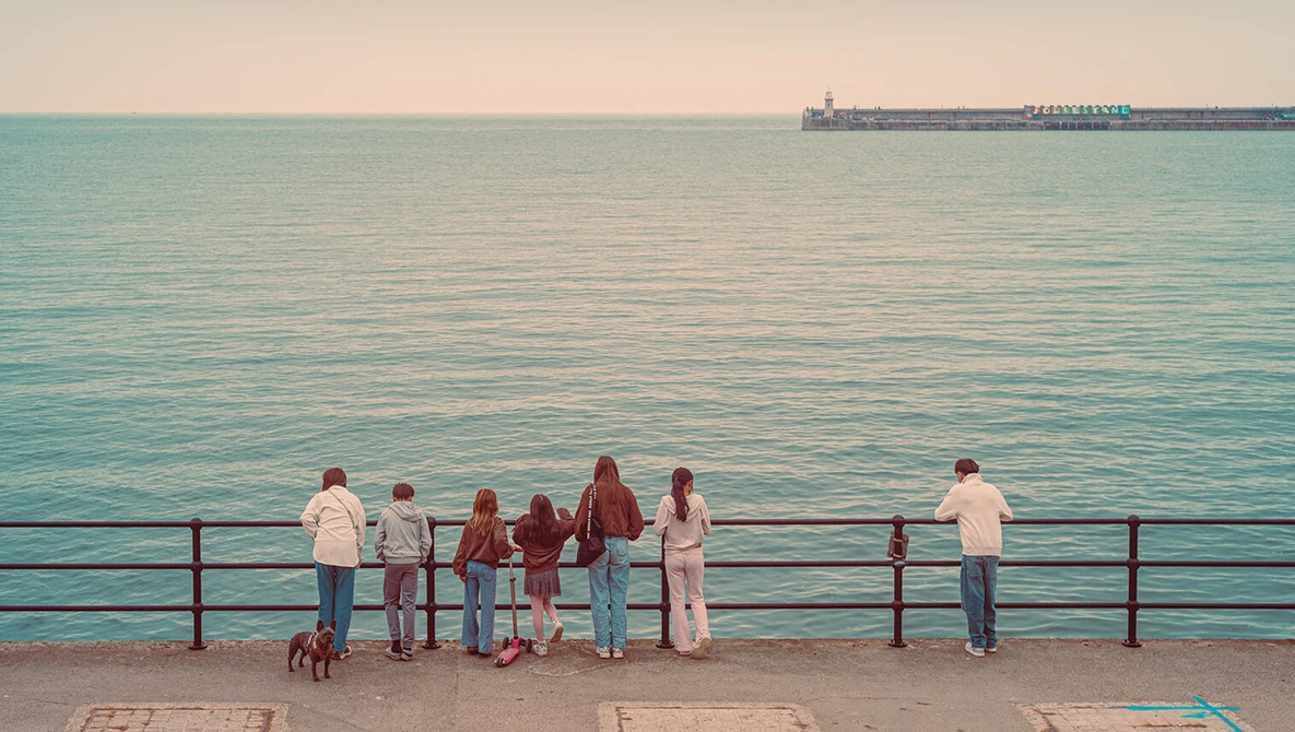 Group of people standing at a seaside promenade railing, overlooking turquoise water with a cargo ship on the horizon