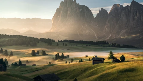 Alpine valley at sunrise with dramatic peaked mountains, mist-filled meadows, and scattered wooden buildings.