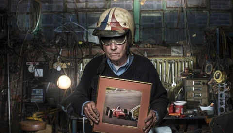 Craftsman in a workshop holding a framed photograph of a race car, surrounded by hanging tools and equipment.