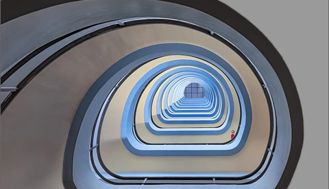Overhead view of a spiral staircase descending through concentric rounded rectangular frames in blue, beige, and gray tones.