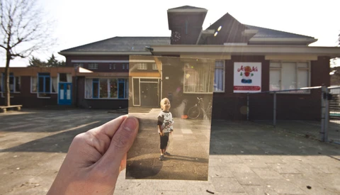 Hand holding a photograph showing a child standing in a schoolyard, with the school building visible in the background