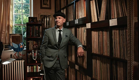 Man in gray suit and fedora standing in a library lined with vinyl records and books.