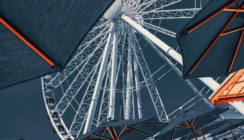 Upward view of a Ferris wheel's intricate white metal framework against a dark blue sky, with orange structural supports visible on either side.