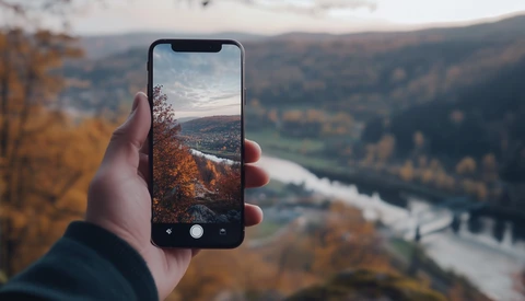 Hand holding smartphone capturing autumn valley landscape with river winding through forested hills.