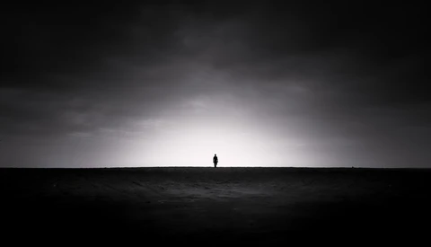 Silhouetted figure standing alone on a dark horizon beneath a dramatic cloudy sky with bright light breaking through.