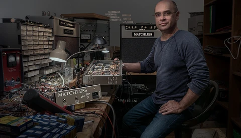Man in home electronics workshop holding circuit board with soldering equipment visible.