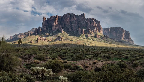 Desert landscape with dramatic rocky butte and saguaro cacti under partly cloudy sky.