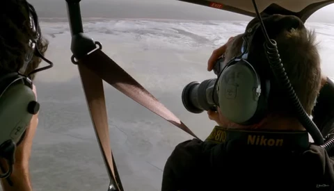 Aerial photographer with Nikon camera shooting from a helicopter, capturing landscape below through open door.