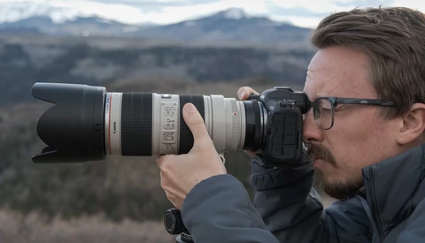 Photographer with glasses holding a telephoto lens camera, shooting toward distant snow-capped mountains.