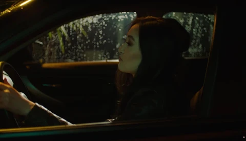 Woman in dark clothing seated in car at night, looking out window with rain-covered glass.