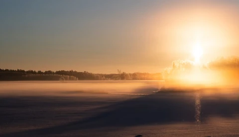 Golden sunrise over a frozen lake with forested shoreline and mist rising from the water.