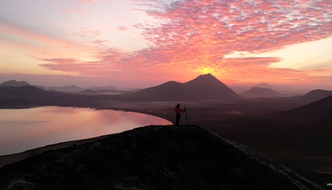 Silhouetted figure standing on a dark ridge overlooking a lake at sunset with volcanic peaks in the distance.