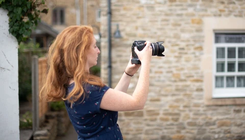 Woman with reddish-blonde hair holding a camera up to her face while standing in front of a stone building.